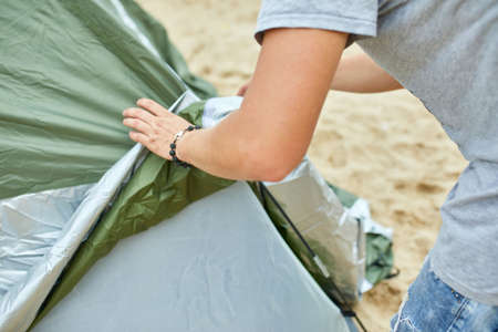 Young male tourist puts a green tent in the beach coast . The man sets up a camp on a hike, collects a tent alone, new normal travel.の写真素材