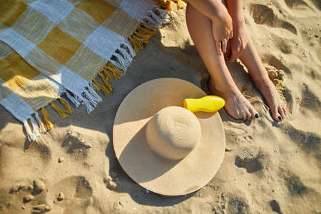 Top view Woman leg sitting on the sandy beach, near hat and sunscreen, sunburning sunblock in hot...