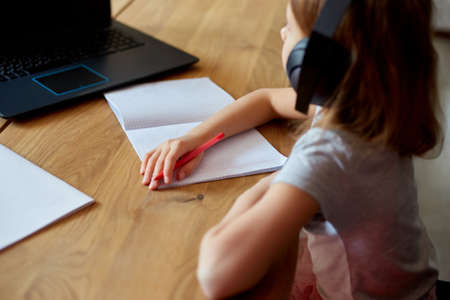 Cute little girl with headphones using laptop to study at home, writing, answer, online learning, education, social distance, quarantine coronavirus.の写真素材