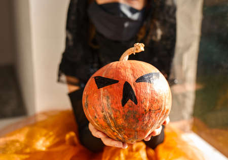 Girl sitting at the window at home in halloween costume with pumpkin Jack or Laurent, Child wearing black face mask protecting from virus, halloween in quarantine.の写真素材