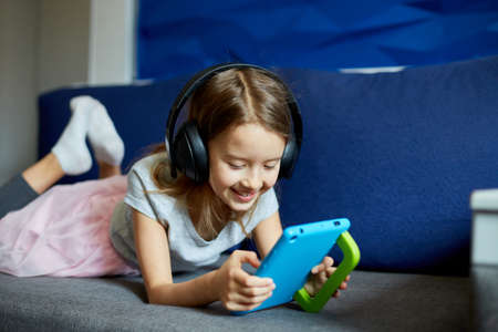 Cute little girl in headphones lying on couch with on digital tablet computer, Kid addicted to technology, enjoying playing online game, using applications, web surfing information at home.の写真素材
