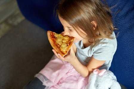Cute little girl sitting on couch and eating piece of italian Pizza at home, yummy mealの写真素材