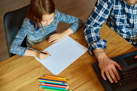 Top view of Father working in his home office on a laptop, her daughter sits next to her and draw,...