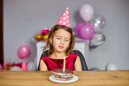 A little girl is wearing a birthday hat makes a wish , looking at a Birthday Cake, with glowing candles for a celebration party concept.の写真素材