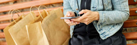 Banner of Woman sitting on the bench in the park withpaper shopping bags after shopping, holding in hands smartphone, typing text message via cellphone, charting in mobile phone. Black Fridayの写真素材