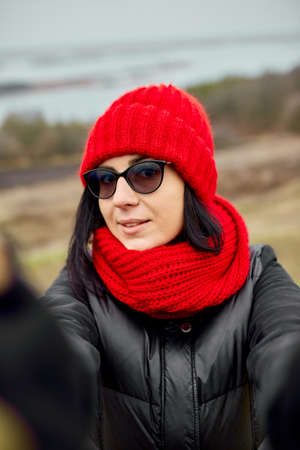 Woman traveler takes a selfie on her phone, wearing a black jacket and a red hat and scarf, autumn background, local travel.の写真素材