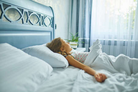 Little girl wakes up from sleep on a big and cozy bed white linen in the at home, family lifestyle,...