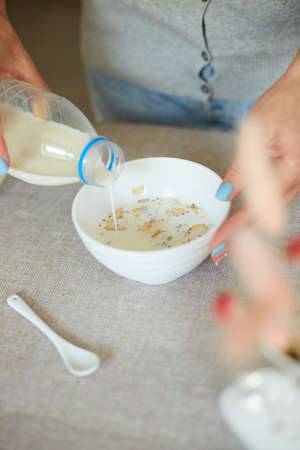Woman Preparing Healthy Breakfast At Home, Female hand holding bottle pouring milk in cereal granola flakes bowl with nuts seeds raisins, home muesli food oat meal, lifestyle conceptの写真素材