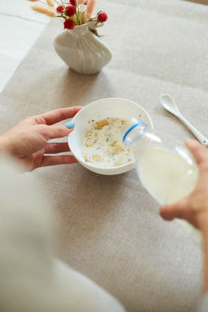 Woman Preparing Healthy Breakfast At Home, Female hand holding bottle pouring milk in cereal granola flakes bowl with nuts seeds raisins, home muesli food oat meal, lifestyle conceptの写真素材