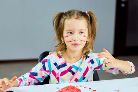 Cute child sitting at the table and drawing stripe camouflage on her face by hand, Art school concept, girl kid painting with watercolor paintsの写真素材
