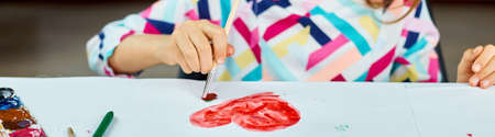 Banner Cute child sitting at the table and drawing red heart on white paper, Art school concept, girl kid painting with watercolor paints at homeの写真素材