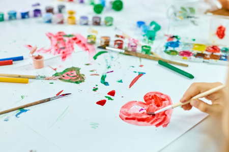 Cute child sitting at the table and drawing red heart on white paper, Art school concept, girl kid painting with watercolor paints at homeの写真素材