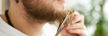 Banner Close up of Handsome man in white t-shirts cutting beard, moustache personally himself with scissors at home. Self Care during quarantine isolation. Stay home.の写真素材