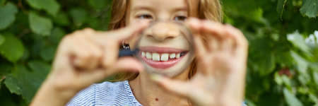 Little girl playing with a magnifying glass in her backyard at home make faces. Childhood lifestyle...