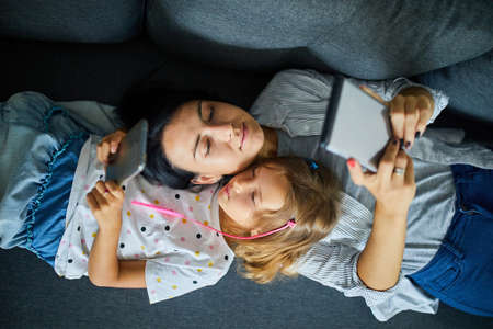 Mother and daughter browsing mobile phone while relaxing together on sofa at home, woman and girl...