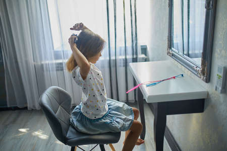 Pretty Little girl combing by herself hair in front of a mirror, brushing her hair at home, child...