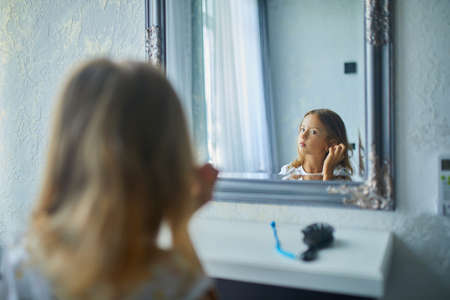 Pretty Little girl combing by herself hair in front of a mirror, brushing her hair at home, child...