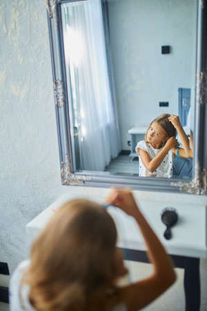Pretty Little girl combing by herself hair in front of a mirror, brushing her hair at home, child smiling, morning routineの写真素材