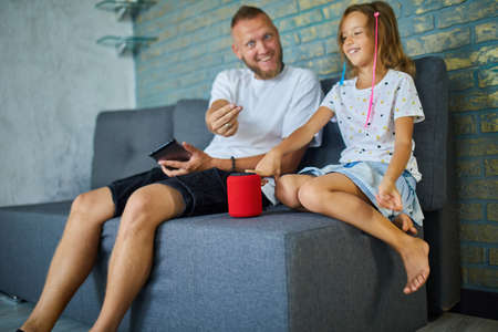 Father and daughter testing talking smart wireless speaker, controlling home devices with a voice commands, sitting on sofa at home, Concept of smart homeの写真素材