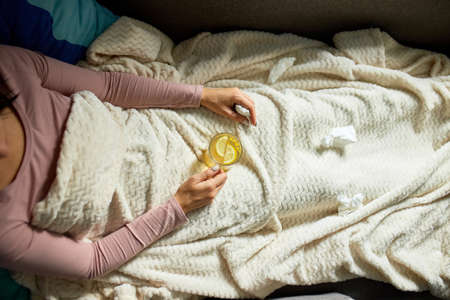 Top view Sick woman holding mug of warm sea buckthorn tea and sitting under blanket at home, Ill...