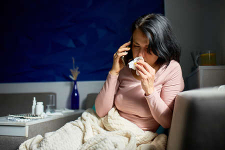 Ill lady wiping nose and measuring temperature at home, talking the phone with doctor. Sick young woman.の写真素材