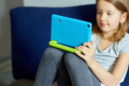 Cute little girl sitting on couch, Kid addicted to technology, enjoying playing online game on digital tablet computer, using applications, web surfing information at home,の写真素材