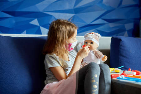 A little girl in medical mask sits on the sofa, plays a doctor, treats and listens to the doll with a stethoscope in a medical mask, at home during quarantine and pandemicの写真素材