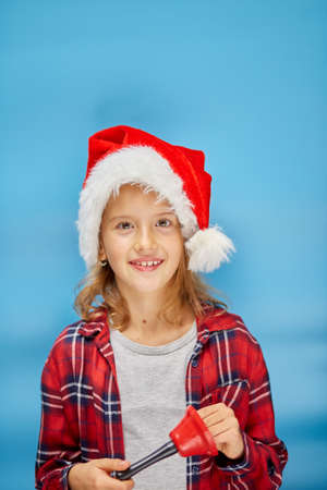Portrait of Christmas little Girl in Santa Hat with bell, smiling child on isolated blue studio...