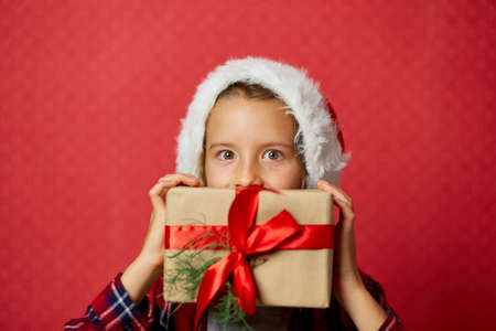 Cute little girl in santa hat holding Christmas gifts in front of her face on isolated red studio background, place for textの写真素材