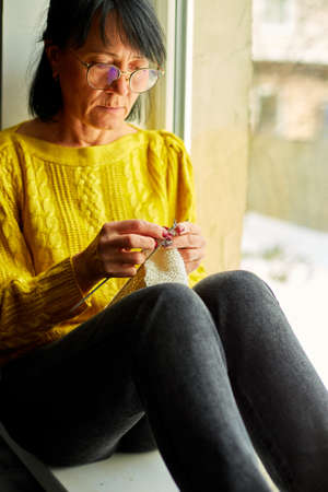 Mature female sitting on the windowsill and knitting while enjoying weekend at home, Senior woman knitting, relax at home do favorite hobby activity on weekend, wellbeing conceptの写真素材