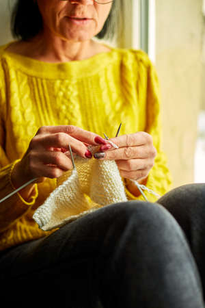 Mature female sitting on the windowsill and knitting while enjoying weekend at home, Senior woman knitting, relax at home do favorite hobby activity on weekend, wellbeing conceptの写真素材