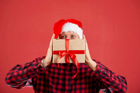 Man in santa hat holding Christmas gifts in front of his face on isolated red studio background, place for textの写真素材