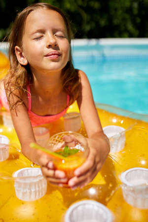 Little girl relaxing in swimming pool, enjoying suntans, drink a juice on inflatable yellow mattress...