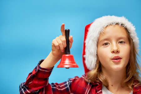 Portrait of Christmas little Girl in Santa Hat with bell, smiling child on isolated blue studio background, place for textの写真素材