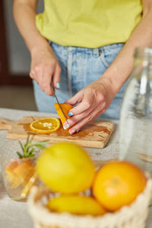 Female hand cut orange, Woman preparing, making citrus and rosemary fresh lemonade in glass on a white table at home, summer drink, detox healthy water.の写真素材