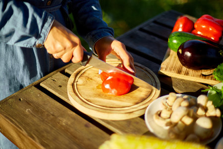 Unrecognizable woman cutting fresh pepper vegetables on wooden board during weekend barbecue in...