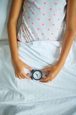 Little girl hold in hand an alarm clock, wakes up from sleep on a big and cozy bed white linen in the at home, family lifestyle, comfortable pillow, bed, Good morning, time to school.の写真素材