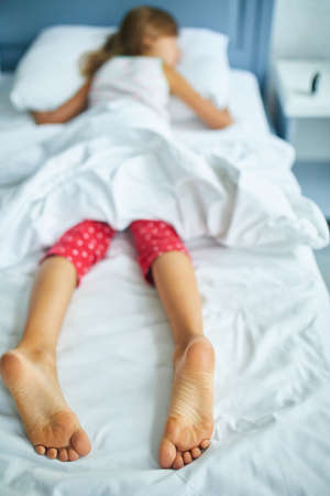 Girl's feet covered with white bed sheet, sleeping in a comfortable bed, Focus on the foot, healthy sleep of childrenの写真素材