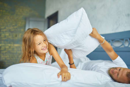 Happy daughter and father playing pillow fight in bed at morning at home, fighting with pillows, have fun play together at home on family weekendの写真素材
