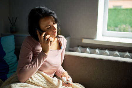 Ill lady measuring temperature at home, talking the phone with doctor. Sick young woman.の写真素材