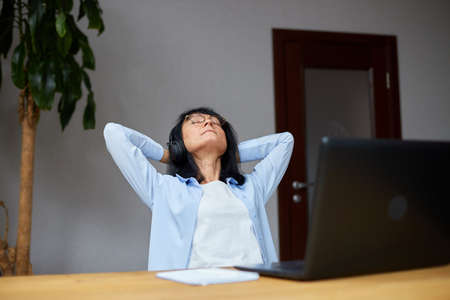 Business mature woman relaxing wearing headphone and listening music, podcast with closed eyes relaxing on the break, sitting at the workplace at home. Elderly ladyの写真素材