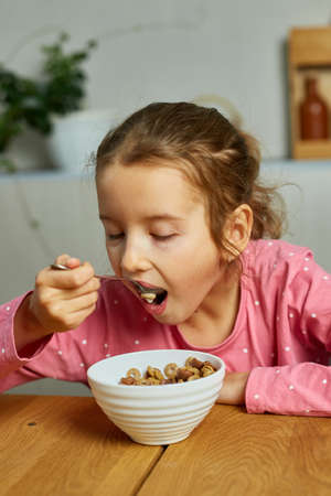 Cute little girl enjoy eating cereal with milk for morning breakfast with appetite, How tasty healthy food, Slow motion of beautiful child having breakfast at home.の写真素材