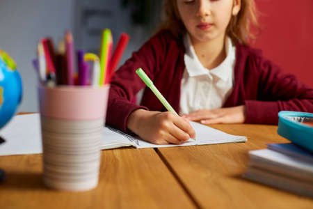 Focused schoolgirl sit at desk doing homework handwriting, homeschooling. Small junior girl engaged in distant learning get remote elementary education and preparing for exams.の写真素材