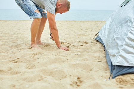 Young male tourist puts a green tent in the beach coast . The man sets up a camp on a hike, collects a tent alone, new normal travel.の写真素材