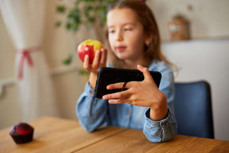 Beautiful young girl filming her blog broadcast about healthy food at the home. Little girl choosing between sweet cake and fresh red apple in , Healthy dieting habit, concept of proper nutritionの写真素材
