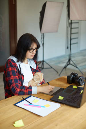 Creative female photographer with cute cat, using graphic drawing tablet and stylus pen, working at desk and retouch photo on tablet computer, Retoucher workplace in photo studio Home office with petの写真素材
