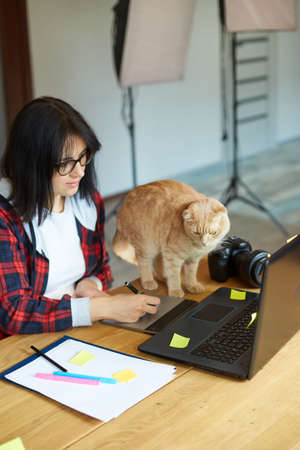 Creative female photographer with cute cat, using graphic drawing tablet and stylus pen, working at desk and retouch photo on tablet computer, Retoucher workplace in photo studio Home office with petの写真素材