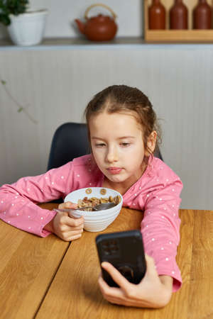 Little girl eating cereal with milk and watching video on smartphone, tasty healthy food, Slow motion of beautiful child having breakfast at home, dependence from gadgetの写真素材
