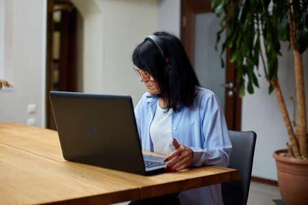Mature woman freelancer in eyeglasses holding meeting via video call from home, make notes, senior...