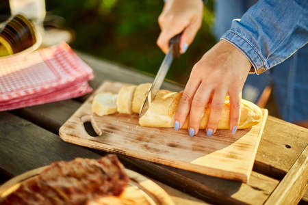 Unrecognizable woman cutting bread for bruschetta on wooden board during weekend barbecue in yard,...